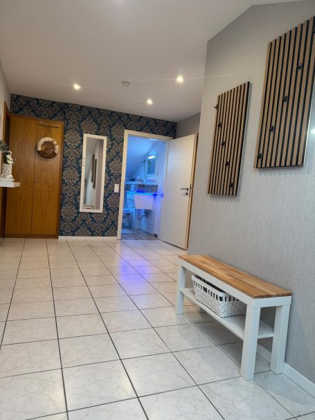 Heller Flur mit Fliesen, Holzbank und dekorativer Wandgestaltung. Tür führt in einen Raum.