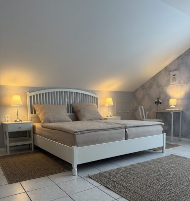 Helles Schlafzimmer mit Doppelbett, Nachttischen und Schreibtisch unter einem schrägen Dach.